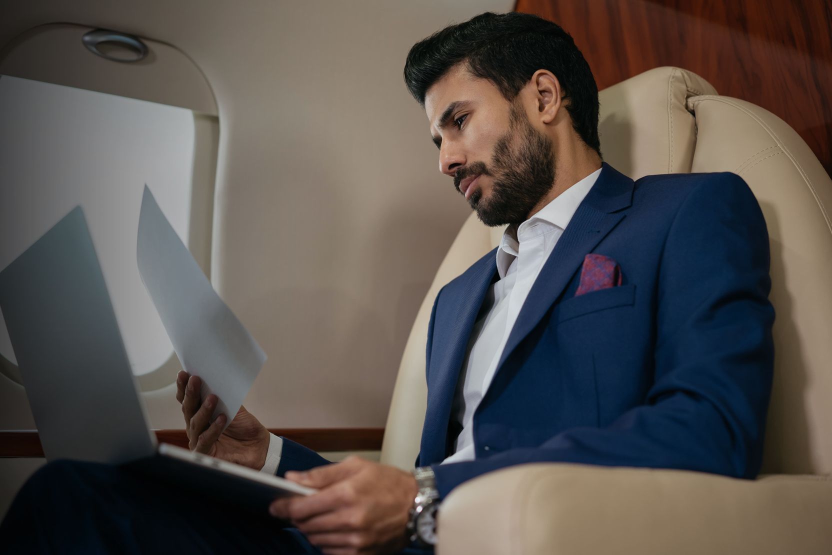 A successful businessman in a blue suit reviewing documents and working on his laptop inside a private jet.