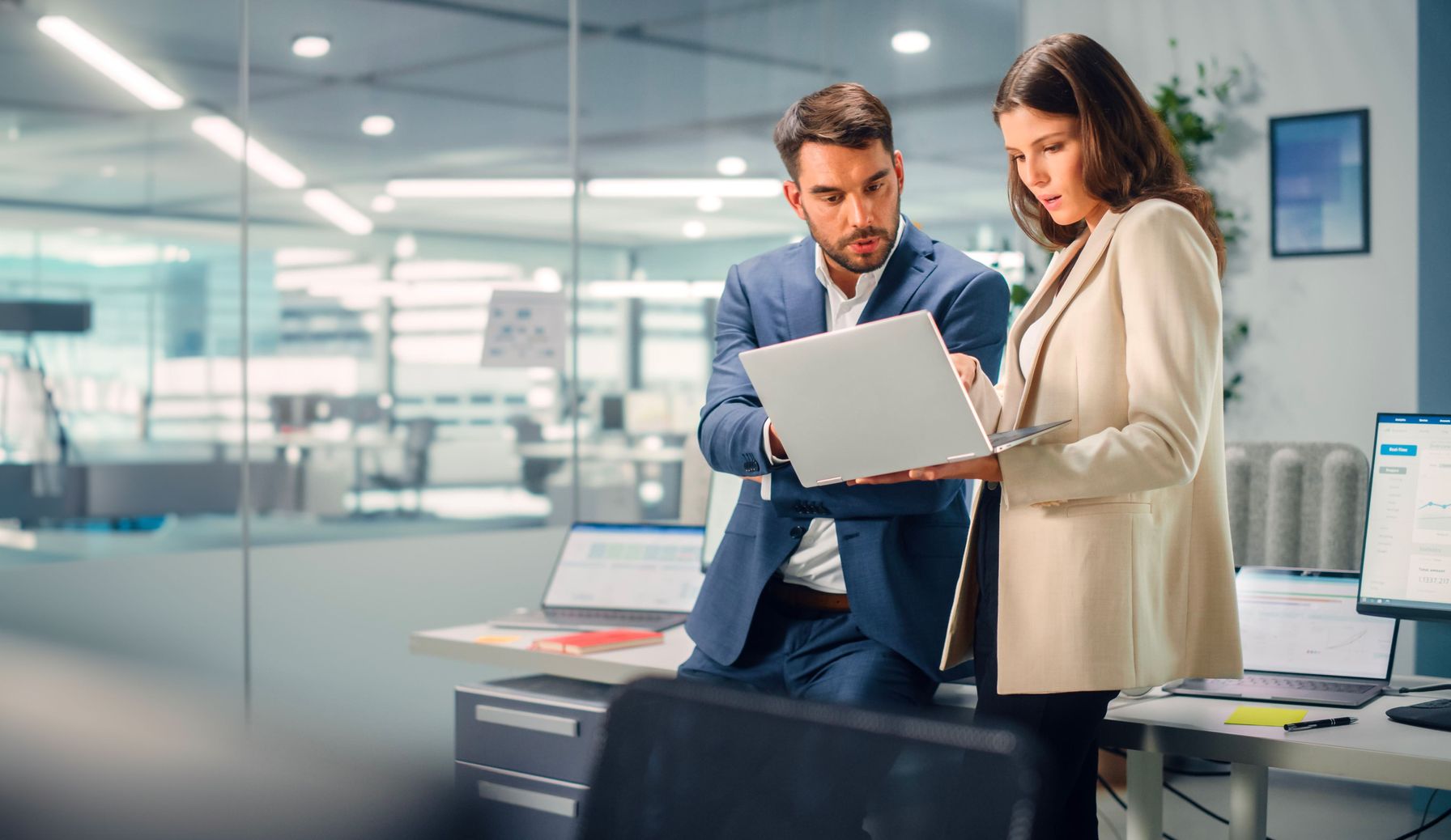 A male and female professional discussing data on a laptop in a high-tech office.