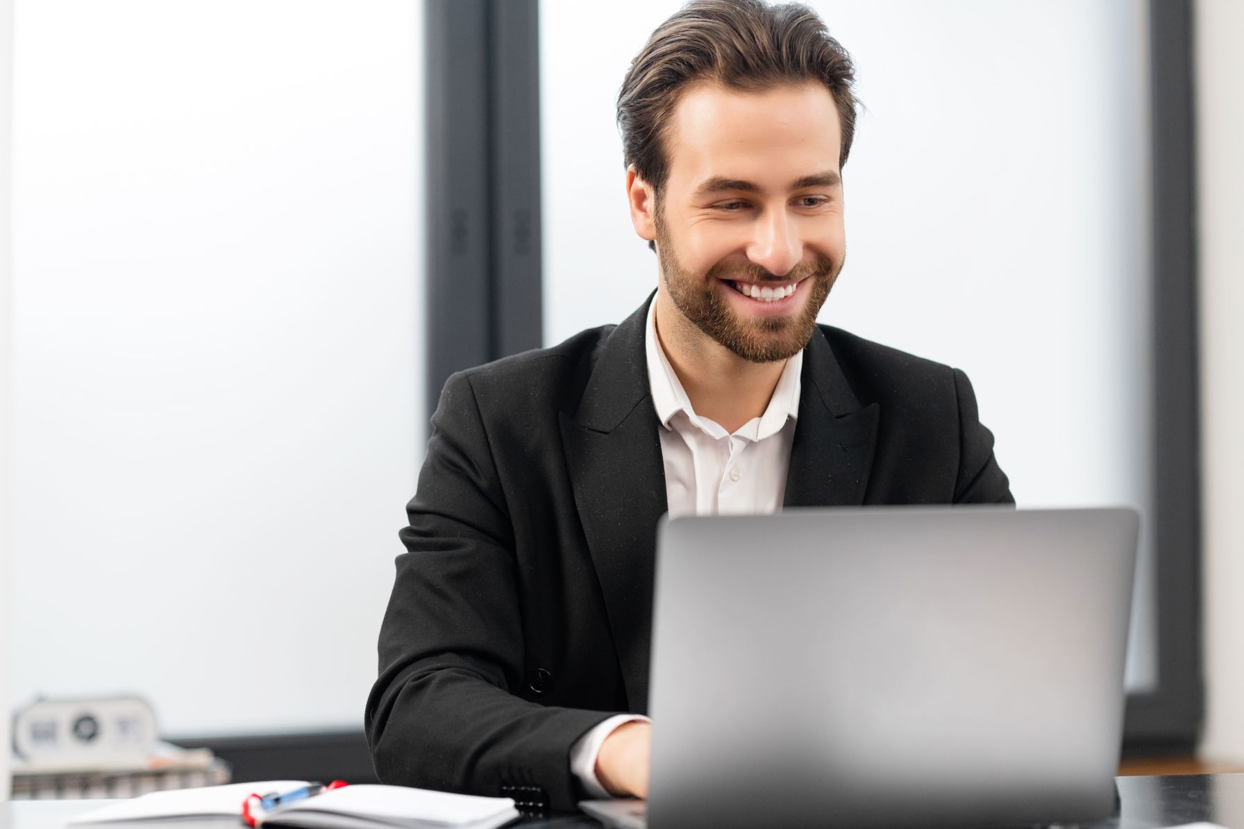 A cheerful businessman in a black suit working on his laptop in a modern office. 