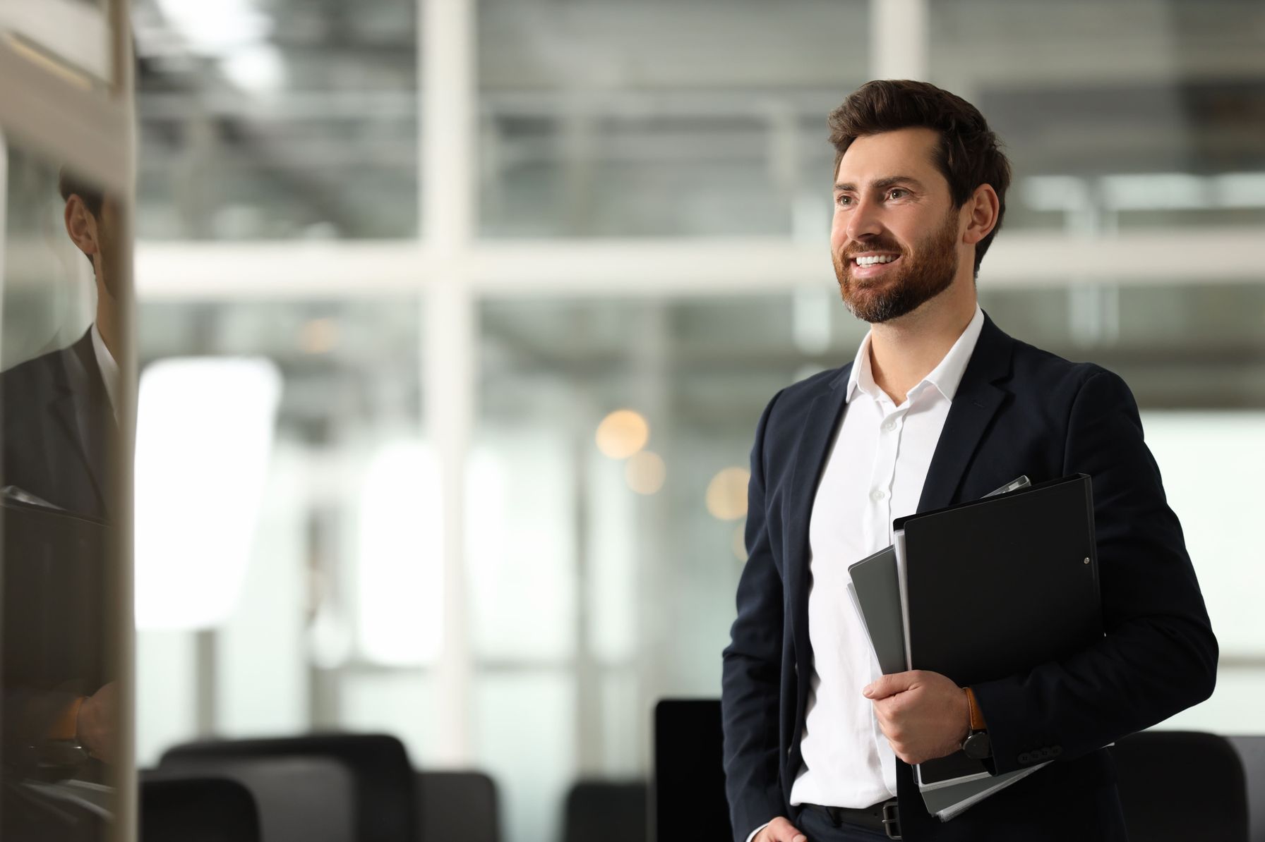A smiling businessman in a formal suit holding documents and a tablet, standing in a bright, modern office.