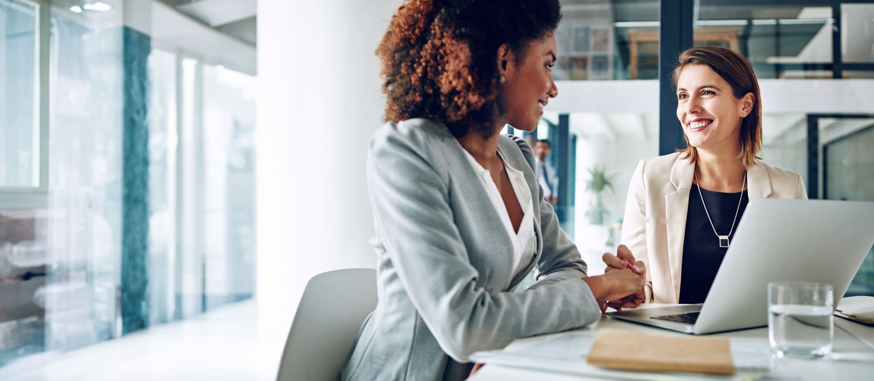 Two professional women in a modern office having a positive conversation while working on a laptop,
