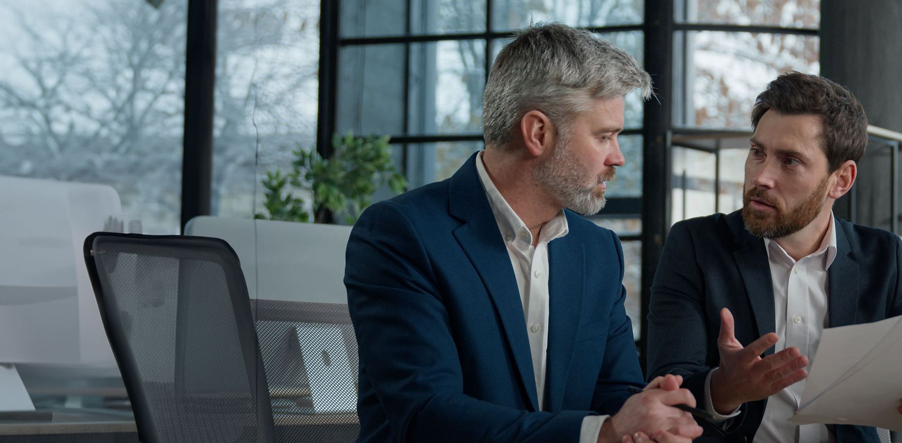 Two businessmen in formal suits engaged in an intense discussion over documents in a modern office.