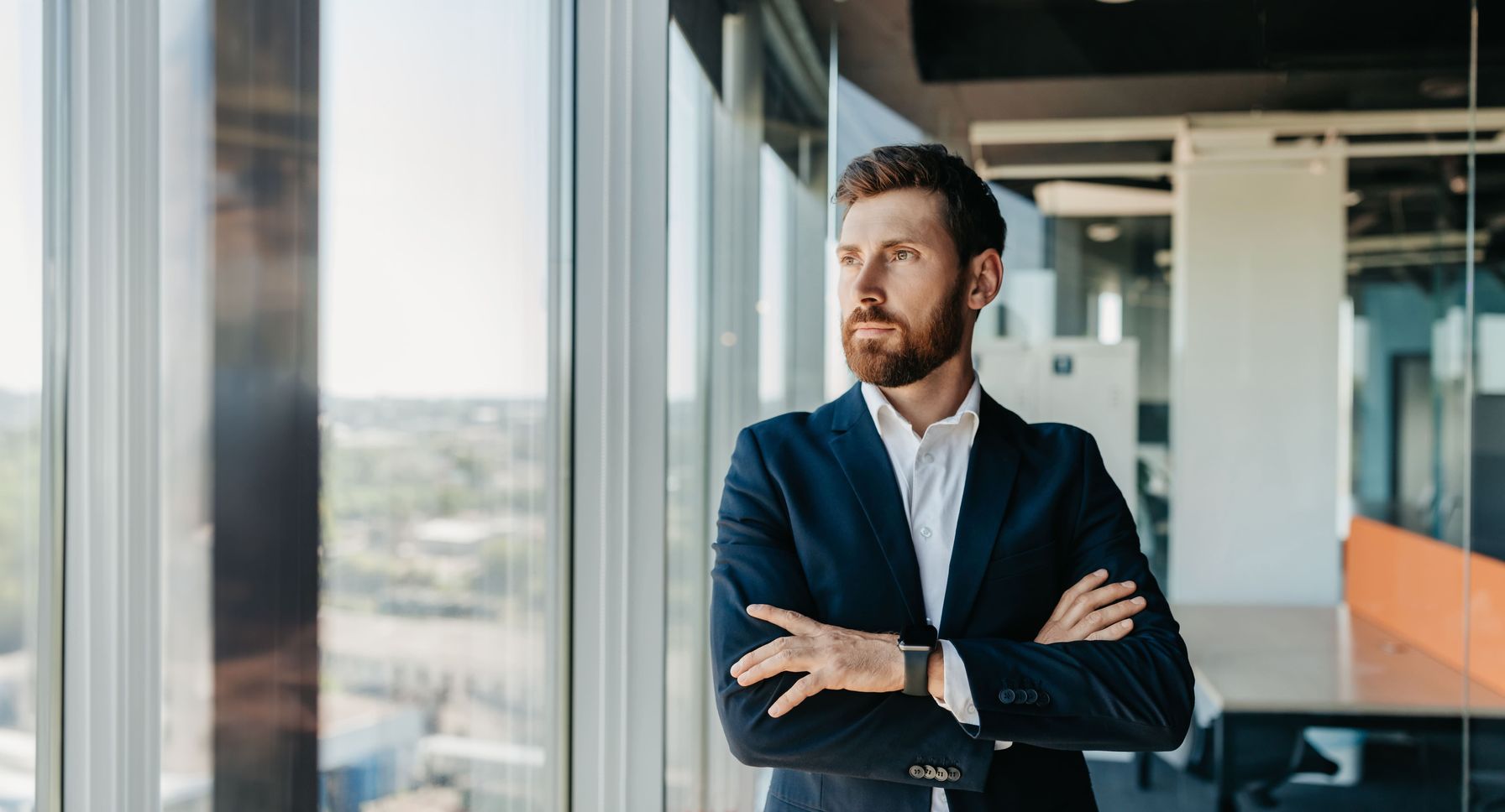 A professional businessman in a navy suit standing with folded arms, gazing out the window in a modern office