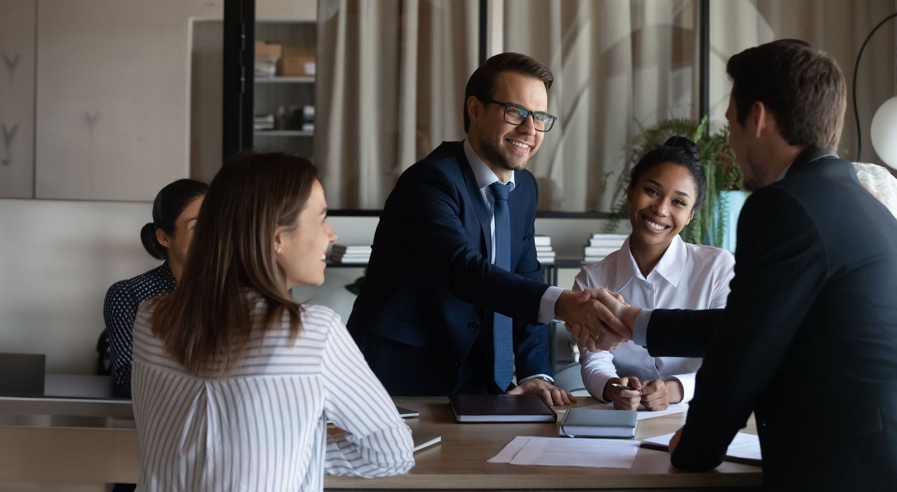 A smiling group of professionals in a modern office celebrating a successful business deal with a handshake.