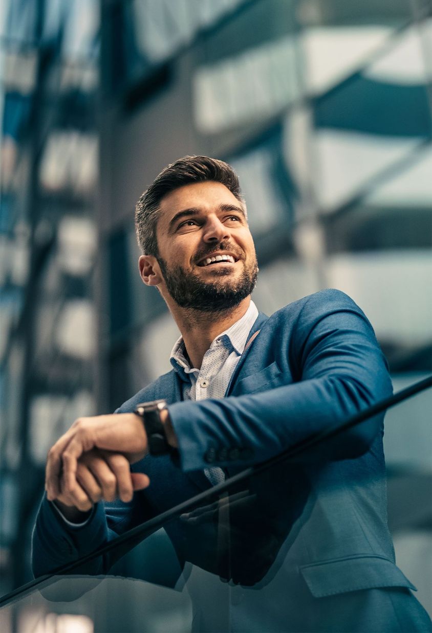 A smiling businessman in a blue suit leaning on a glass railing, surrounded by modern glass architecture.