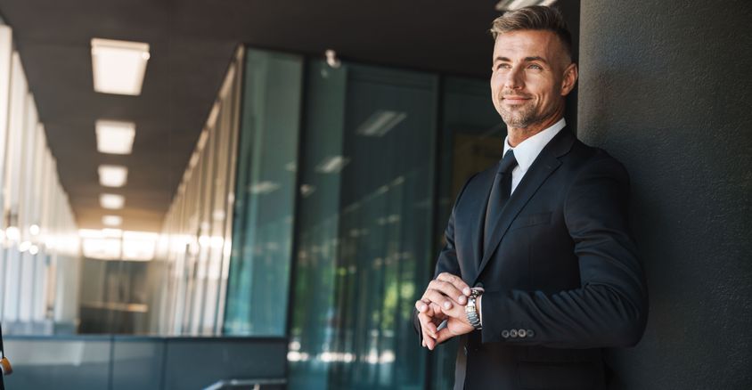 A mature, successful businessman in a black suit standing outside a modern office building, checking his watch with a confident smile.