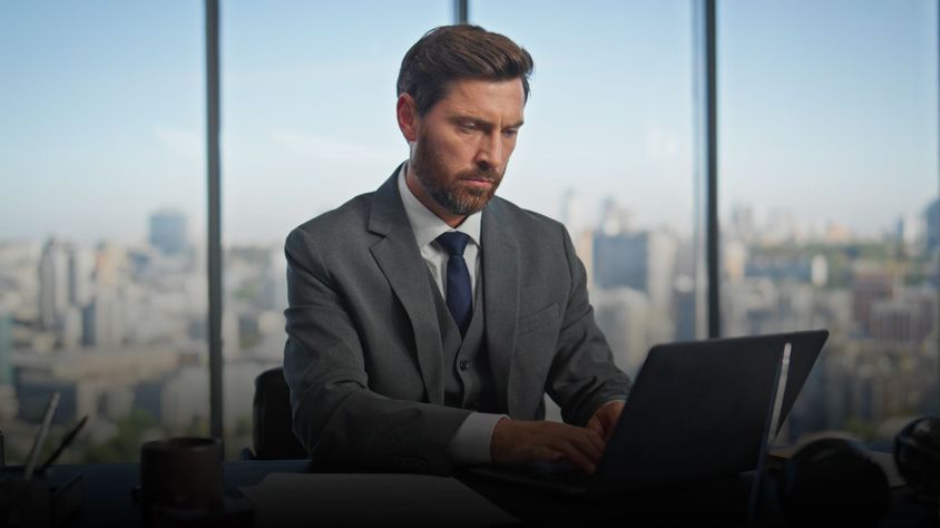 Professional businessman in a suit analyzing data on a laptop in a high-rise office with city view.